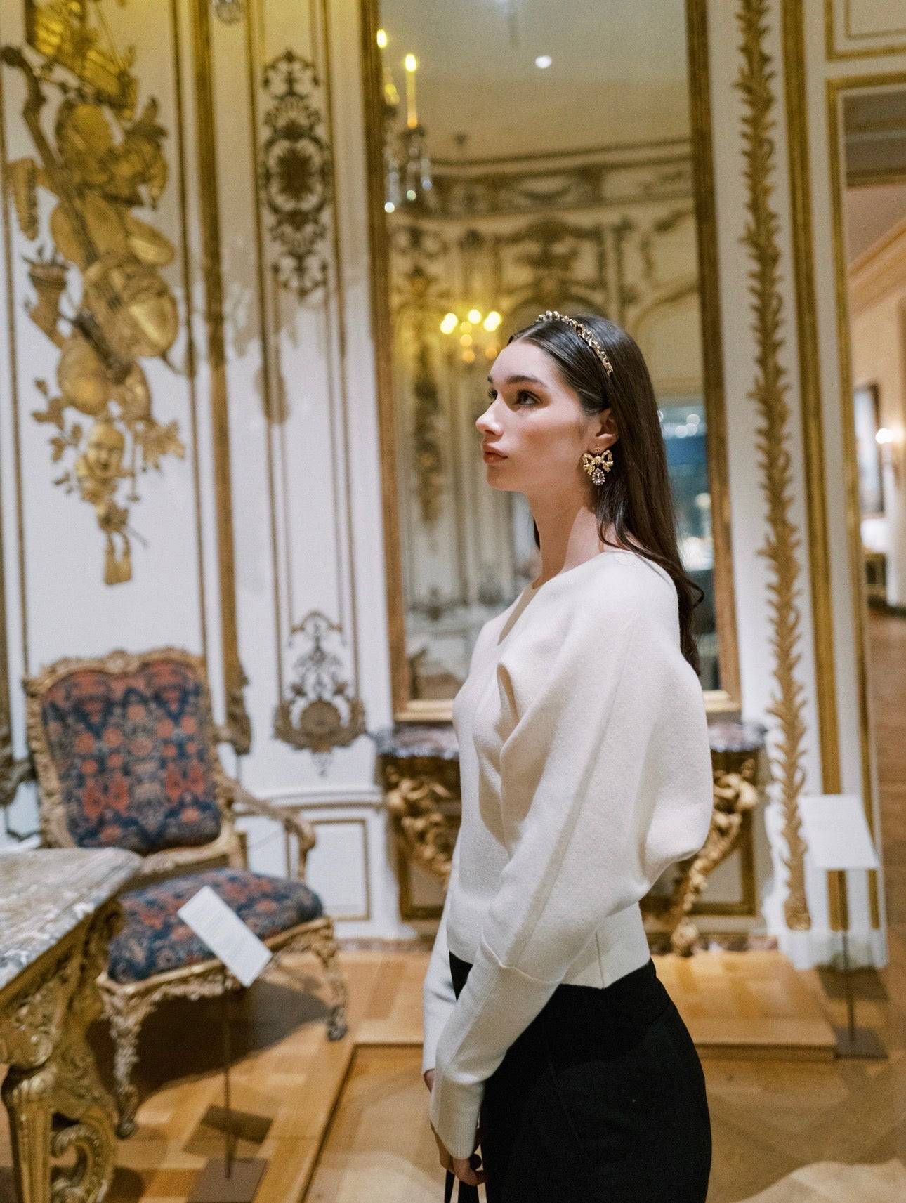 Woman in a white sweater and black pants standing in an ornate room with decorative walls and furniture.