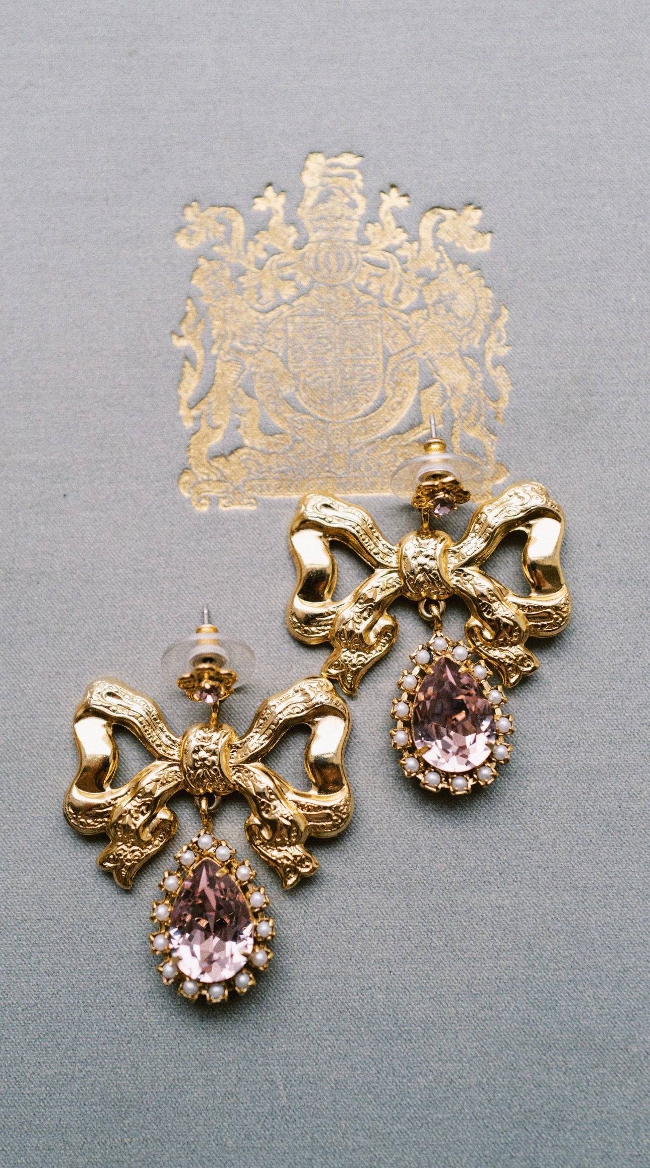 Gold bow-shaped earrings with pink gemstones on a gray background
