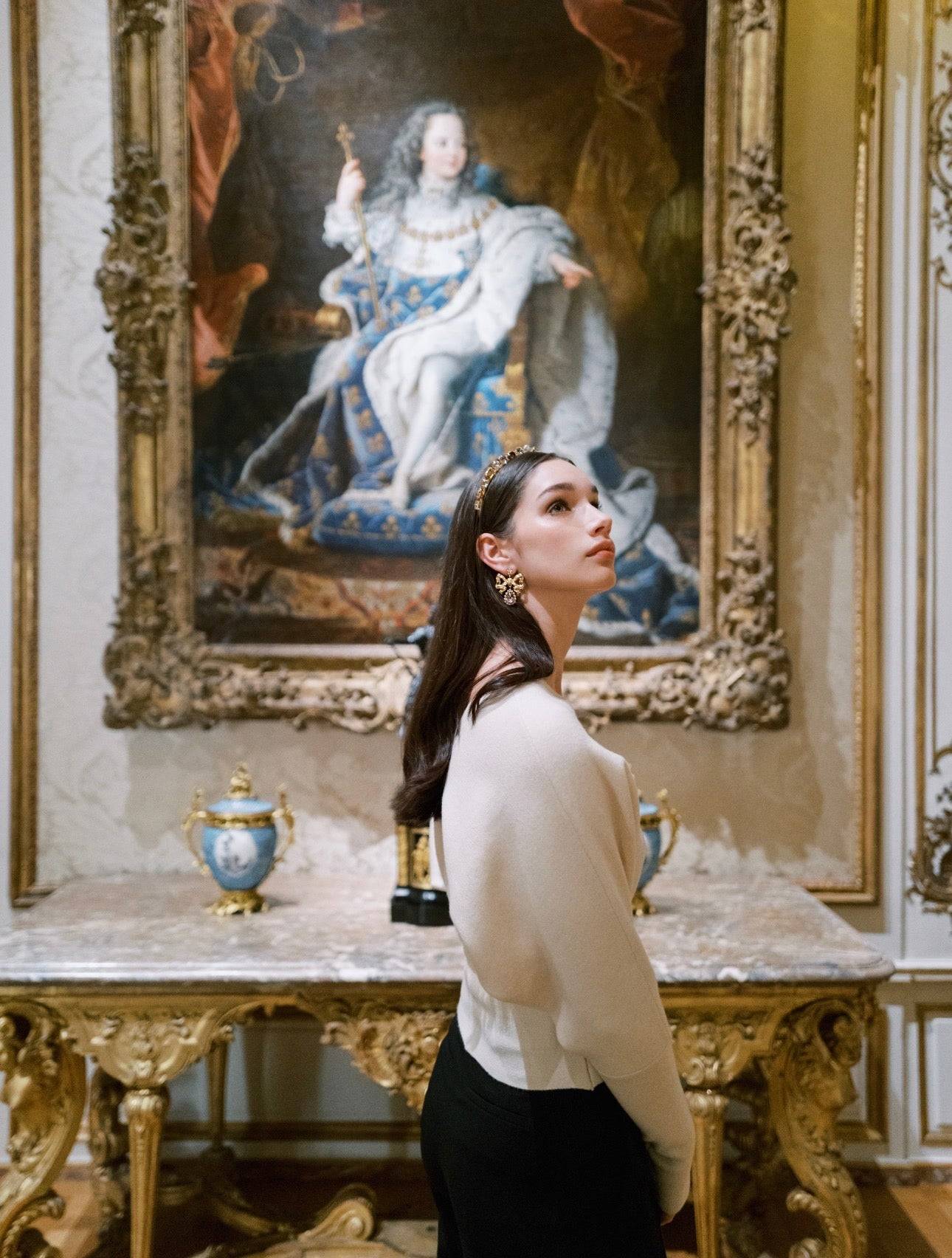 Woman in a white blouse standing in front of a classical painting in an ornate room.