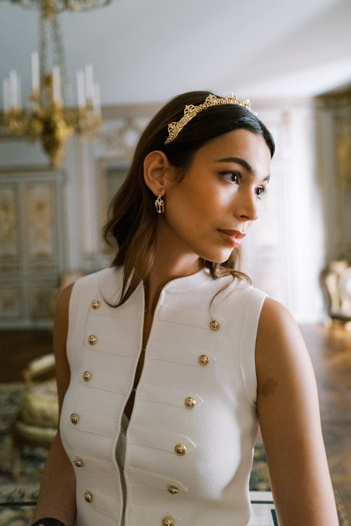 Profile of the Antoinette Halo, a gold baroque scrollwork Swarovski crystal headband, under a chandelier at The Metropolitan Museum of Art, elegant hair accessory for weddings, galas and New Year's Eve | EDEN LUXE Bridal